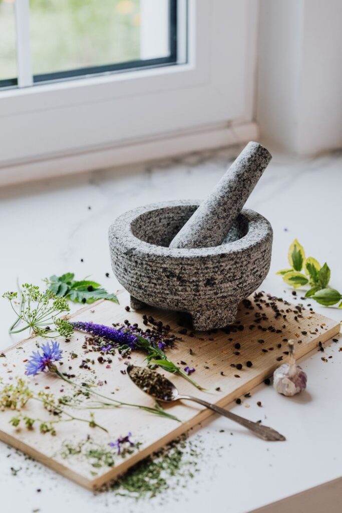 Stone mortar and pestle with scattered herbs and spices on a wooden board near a window.